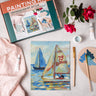 Painting kit with sailboat painting, brushes, and flowers on a table