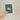 Hand holding a plantable seed paper envelope with a design of two interlocking rings on a green background.