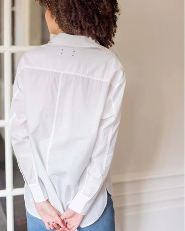 Model is turned away from the camera showing the back of the garment. She is wearing a white button up top and denim pants. The back of the shirt is white and you can see the seam down the middle and across the shoulders. Where the tag is sewn in, red thread is visible at the corners.