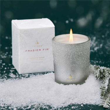 Image shows Thymes Frasier Fir flocked votive candle and its packaging on a deep green background with powdery snow in the foreground.