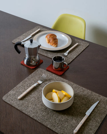 A styled breakfast table setting featuring gray felt placemats, a silver moka pot, a ceramic bowl with sliced fruit, a plate with a croissant, and a small metal cup on a dark wood table with a lime green chair in the background.