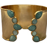 A wide gold cuff bracelet decorated with two vertical columns of teardrop and oval green stones, photographed close up on a neutral surface.