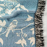 Wren Blanket – Fringe | Chambray. Birds sit among branches in this white on blue design. There is fringe around the edges alternating black/white and multicolored.